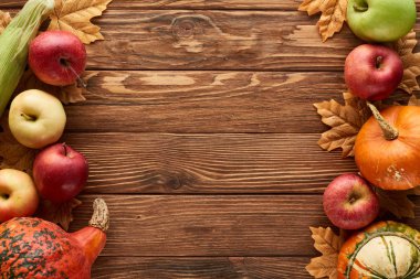 top view of pumpkins, sweet corn and apples on wooden surface with dried autumn leaves clipart
