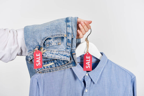 cropped view of woman holding jeans and shirt with sale labels isolated on white