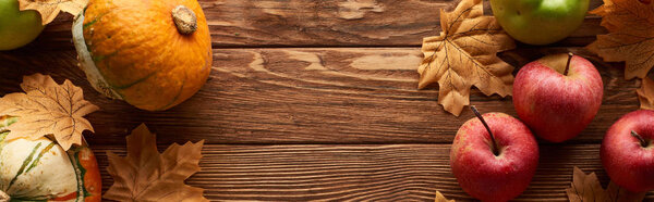 panoramic shot of small pumpkins and apples on brown wooden surface with dried autumn leaves