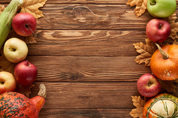 top view of pumpkins, sweet corn and apples on wooden surface with dried autumn leaves