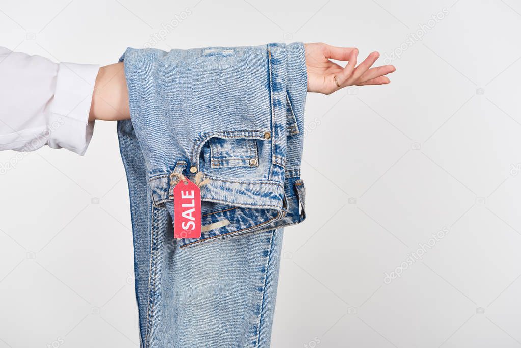 cropped view of woman holding hanging jeans with sale label isolated on white