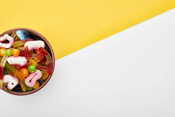 top view of colorful gummy sweets in bowl on yellow and white background, Halloween treat