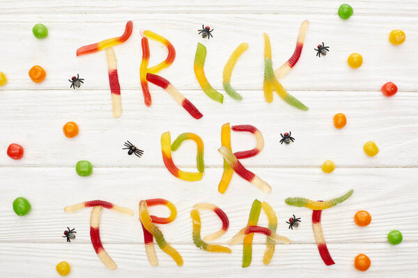 top view of trick or treat lettering made of colorful gummy sweets on white wooden table with spiders and bonbons, Halloween treat