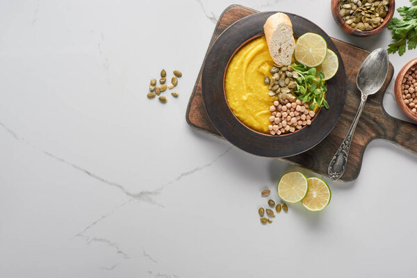 top view of autumnal mashed pumpkin soup in bowl on wooden cutting board with spoon on marble surface