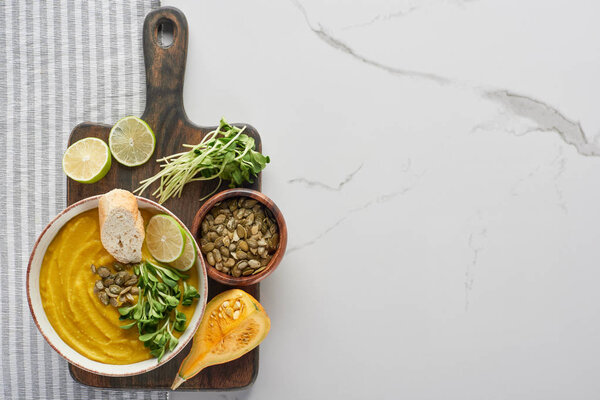 top view of tasty mashed pumpkin soup on wooden cutting board with ingredients on marble surface
