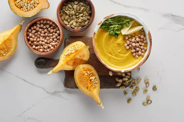Top view of autumnal mashed pumpkin soup in bowl on wooden cutting board near pumpkin, seeds and chickpea on marble surface — Stock Image Top View Autumnal Mashed Pumpkin Soup Bowl Wooden Cutting Board Stock Image