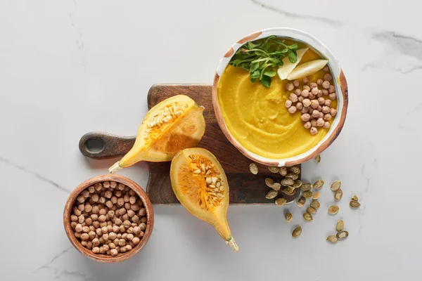 Top view of autumnal mashed pumpkin soup in bowl on wooden cutting board near pumpkin and chickpea on marble surface — Stock Image Top View Autumnal Mashed Pumpkin Soup Bowl Wooden Cutting Board Stock Picture