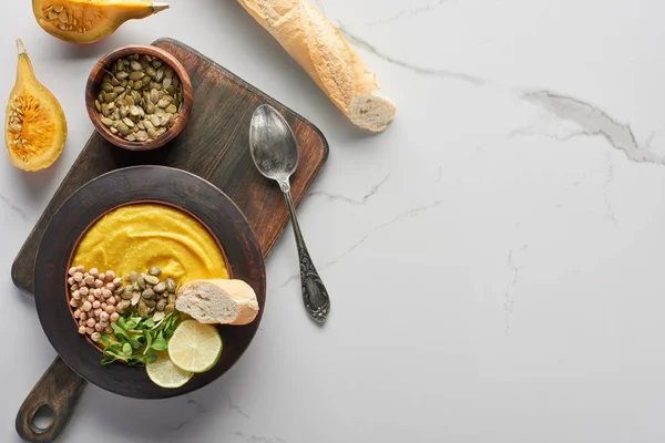 Top view of autumnal mashed pumpkin soup on wooden cutting board near silver spoon and baguette on marble surface — Stock Image Top View Autumnal Mashed Pumpkin Soup Wooden Cutting Board Silver Royalty Free Stock Photos