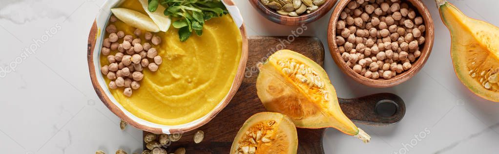 Top view of autumnal mashed pumpkin soup in bowl on wooden cutting board near pumpkin, chickpea on marble surface, panoramic shot — Stock Image Top View Autumnal Mashed Pumpkin Soup Bowl Wooden Cutting Board Royalty Free Stock Images
