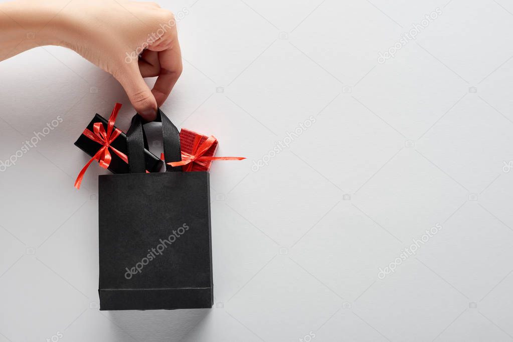 Cropped view of woman holding small black shopping bag with decorative gift boxes on white background