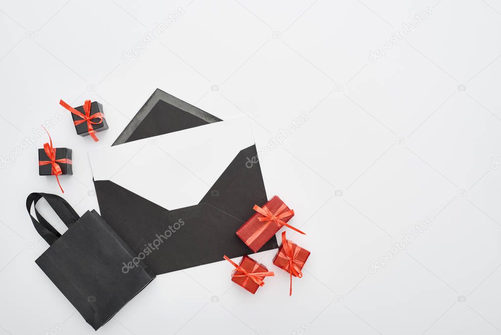 top view of black envelope with blank card near decorative gift boxes and shopping bag on white background