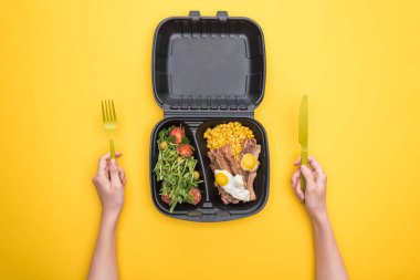 Elinde plastik çatal, bıçak ve ekolojik paket bulunan mısır, et, sahanda yumurta ve salata tutan bir kadın. 