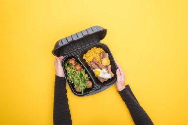 Kırpılmış görüntüde elinde mısır, et, sahanda yumurta ve salata bulunan ekolojik paket olan bir kadın var. 