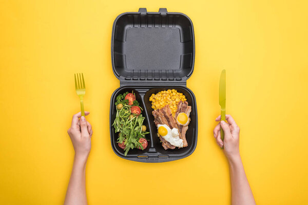 cropped view of woman holding plastic fork, knife and eco package with corn, meat, fried eggs and salad isolated on yellow 