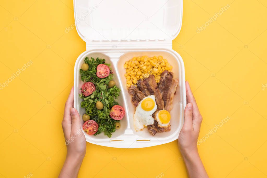 cropped view of woman holding eco package with corn, meat, fried eggs and salad isolated on yellow 