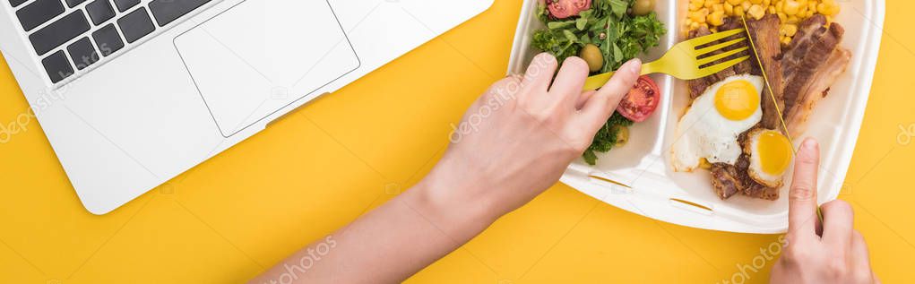 panoramic shot of woman eating from eco package with corn, meat, fried eggs and salad isolated on yellow    