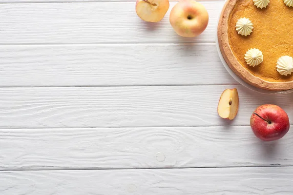カットの近くにホイップクリームと白い木製のテーブルの上にリンゴ全体とおいしいカボチャのパイのトップビュー — ストック写真