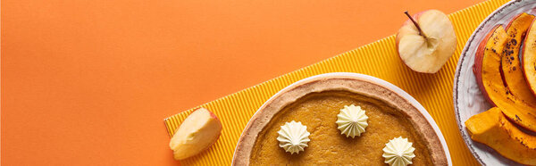 panoramic shot of delicious pumpkin pie with whipped cream near sliced baked pumpkin and cut apple on orange surface