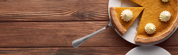 panoramic shot of delicious pumpkin pie with whipped cream on plate with spatula on brown wooden table