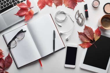 Akıllı telefon, dijital tablet, kahve fincanı, kozmetik, kulaklık, gözlük, defter ve kırmızı üzüm yapraklarının yanındaki dizüstü bilgisayarın beyaz masa üzerindeki görüntüsü.
