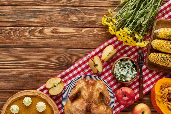 top view of roasted turkey, pumpkin pie and grilled vegetables served on wooden table with flowers and plaid napkin