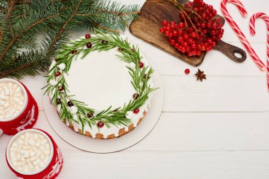 Beyaz ahşap masa üzerinde noel pastası, şeker kamışları, kakao, viburnum üzümleri ve ladin dalları.