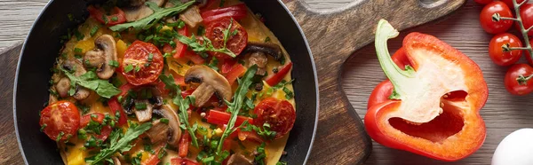 top view of homemade omelet in frying pan with bell pepper and cherry tomatoes on wooden board