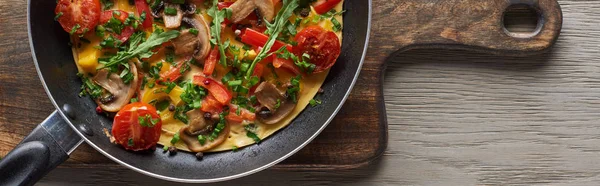 top view of tasty omelet with vegetables and greens in frying pan on wooden board 