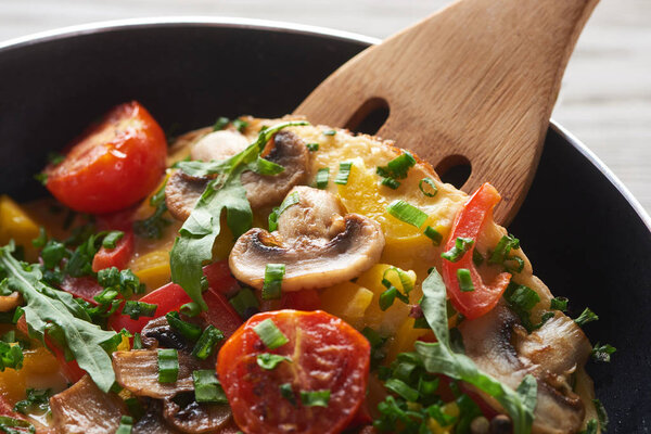 крупным планом домашнего омлета с овощами и зеленью на сковородке с деревянной лопатой
