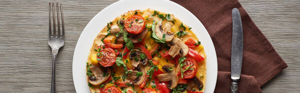 top view of delicious omelet with vegetables on wooden table with fork, knife and napkin