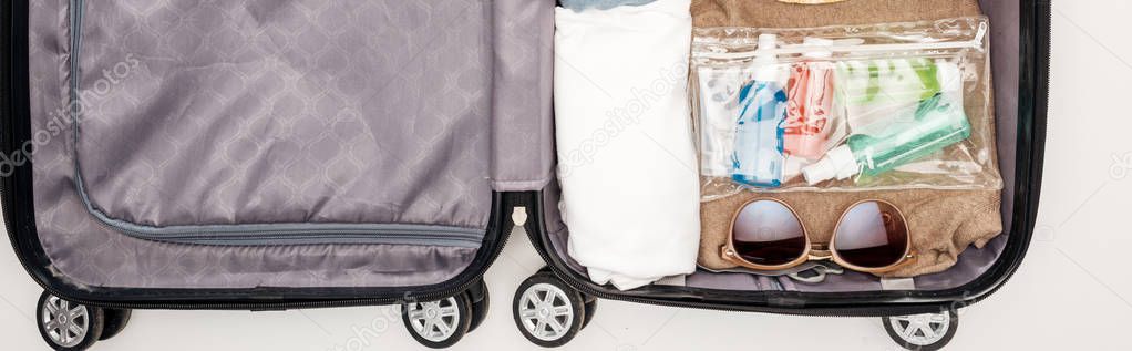panoramic shot of travel bag with towel, cosmetic bag with bottles, clothes, sunglasses on white background 