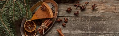 top view of piece of pie, anise, dry orange slices and cinnamon on wooden rustic table near fir branch, panoramic shot clipart