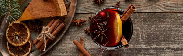 top view of red spiced mulled wine near fir branch, pie, berries, anise, orange slices and cinnamon on wooden rustic table, panoramic shot