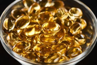 close up view of golden fish oil capsules in glass bowl isolated on black clipart