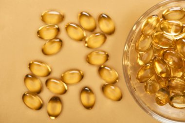 top view of shiny golden fish oil capsules scattered from glass bowl on yellow background clipart