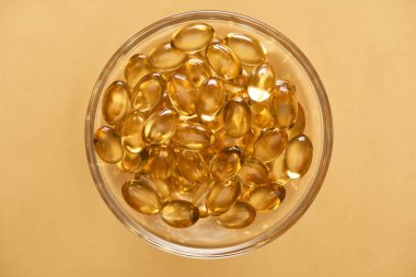 top view of shiny golden fish oil capsules in glass bowl on yellow background clipart
