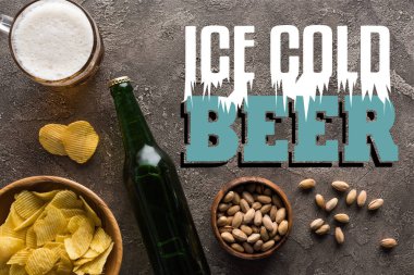 Şişenin yanında şam fıstığı ve cips olan kaselerin üst görüntüsü ve buz gibi bira harfleriyle gri yüzeyde bir kupa bira.