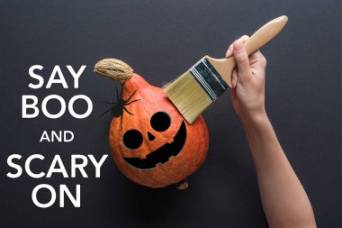 partial view of woman holding paintbrush near pumpkin on black background with say boo and scary on illustration clipart