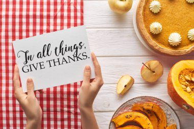 partial view of woman holding card with in all things give thanks illustration near pumpkin pie on wooden white table with apples and plaid red napkin clipart