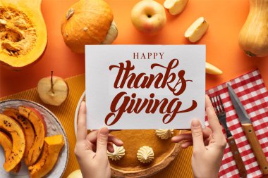 cropped view of woman holding card near with happy thanksgiving illustration pumpkin pie on orange background clipart