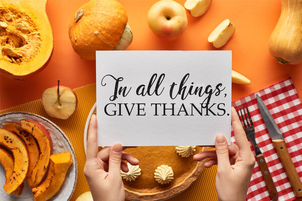 cropped view of woman holding card with in all things give thanks illustration near pumpkin pie on orange background