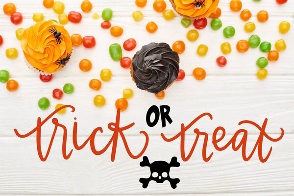 top view of colorful bonbons and cupcakes on white wooden table with trick or treat illustration