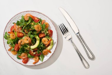 Beyaz arka planda çatal bıçak takımının yanındaki tabakta taze yeşil salata ve avokado var.