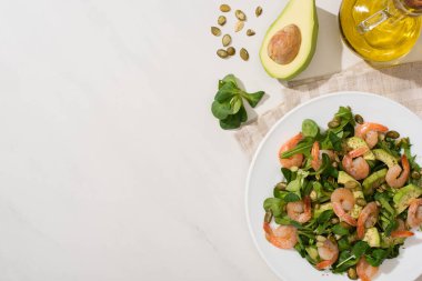 Beyaz arka plandaki malzemelerin yanında kabak çekirdekli taze yeşil salata, karides ve avokado tabakta.