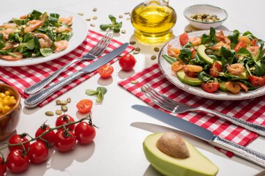 Karidesli taze yeşil salata ve çatal bıçak takımının yanındaki tabakta avokado. Ekose peçeteler ve beyaz masadaki malzemeler.