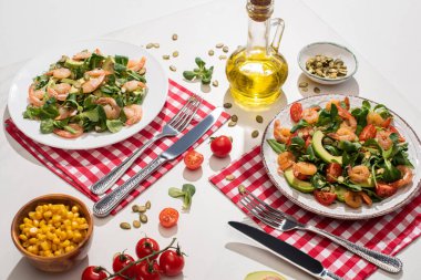 Karidesli taze yeşil salata ve çatal bıçak takımının yanındaki tabakta avokado. Ekose peçeteler ve beyaz masadaki malzemeler.
