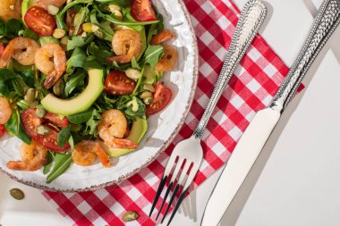 Karidesli taze yeşil salata ve beyaz arka plandaki ekose peçetede çatal bıçak takımının yanında avokado.