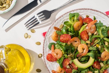 Karidesli taze yeşil salata ve ekose peçetede çatal bıçak takımının yanında avokado ve beyaz arka planda malzemeler.