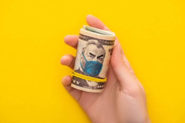 Sarı arka planda tıbbi maskesi çizilmiş para rulosu tutan kadın görüntüsü.