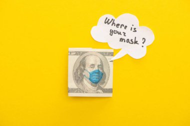 Sarı arka planda tıbbi maskeli ve konuşma baloncuklu dolar banknotunun üst görüntüsü